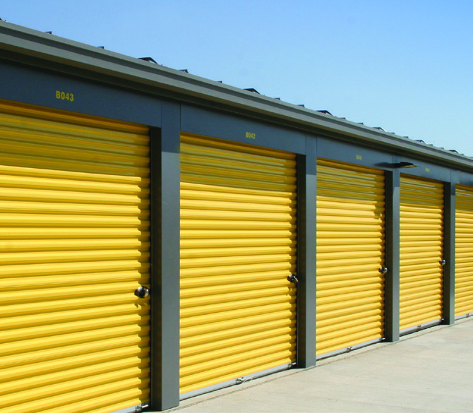 A rows of single-story self-storage units with dark green roll-up doors and a gray metal structural frame, built using a Butler Manufacturing PEMB pre-engineered metal building system.