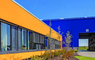 A vibrant industrial building featuring a bright orange office exterior with large glass windows connected to a cobalt blue high-bay warehouse with a large open loading bay.